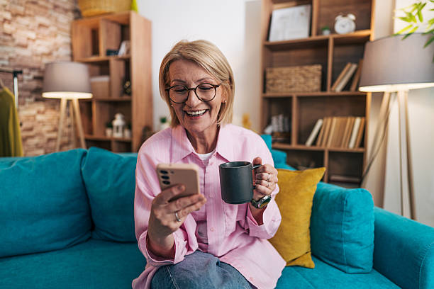 istock Smiling senior woman using smartphone and drinking coffee on sofa at home 2208277258