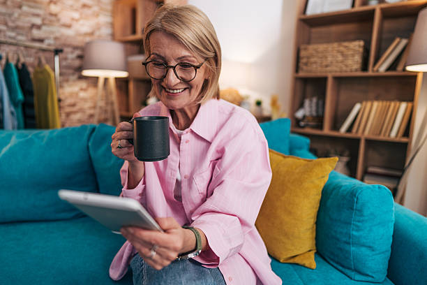 istock Smiling senior woman using digital tablet and drinking coffee on sofa at home 2208277406