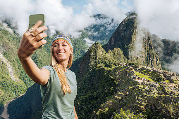 Smiling woman in casual attire capturing selfie with Machu Picchu backdrop. Tourist appreciating scenic landscape. Adventurous travel photography in iconic location.