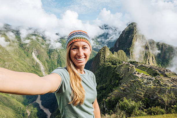 Smiling woman in casual attire capturing selfie with Machu Picchu backdrop. Tourist appreciating scenic landscape. Adventurous travel photography in iconic location.