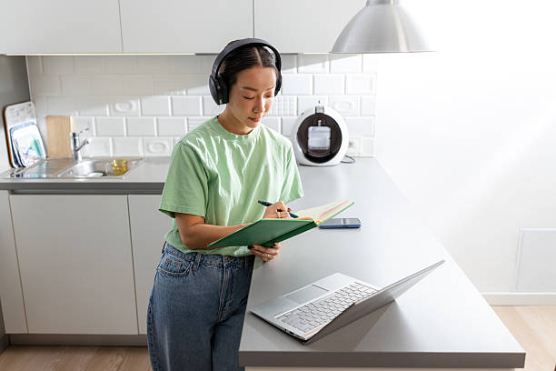 istock Woman with headphones at kitchen counter taking notes while working on laptop 2208282469