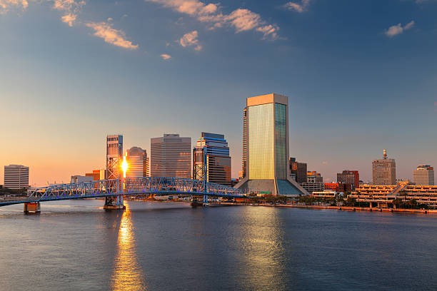 Skyline of Jacksonville, Florida, featuring a mix of modern skyscrapers reflecting golden sunset hues. The prominent blue John T. Alsop Jr. Bridge spans the St. Johns River, connecting both sides. Warm light bathes the buildings, highlighting glass facades and diverse architecture. Sparse clouds accentuate the clear evening sky, creating a tranquil scene. The river reflects the vibrant colors, adding to the city's picturesque charm at dusk.