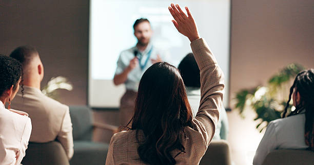 Business people, seminar and convention with audience, question and interaction for feedback or engagement. Group, speaker and back of employee with hand raised for proposal or speech at presentation