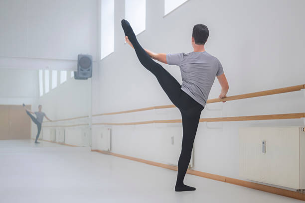A young Caucasian male ballet dancer practices a high leg extension in a well-lit, minimalist dance studio with mirrored walls and wooden bars.