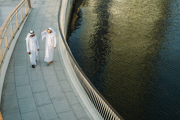 Two Arab businessmen walking and engaging in conversation on a bridge beside a canal, surrounded by Dubai's modern architecture and vibrant city life