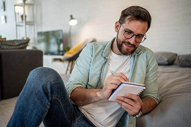 istock Young man sitting on floor and writing in notebook at home 2208311785