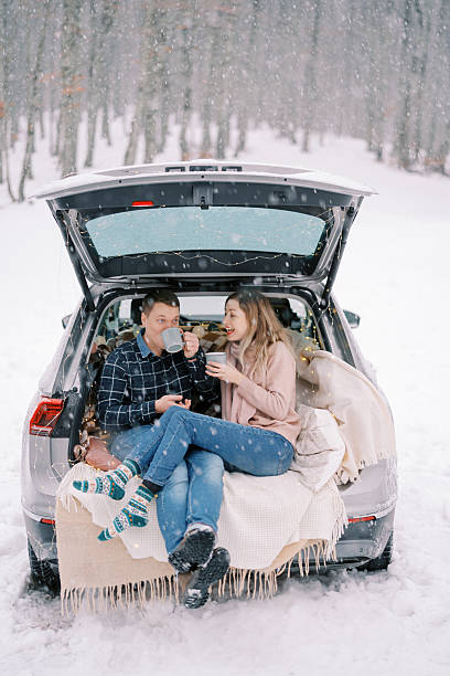 Wife tells something to her husband while he drinks coffee while sitting on blankets in the trunk of a car