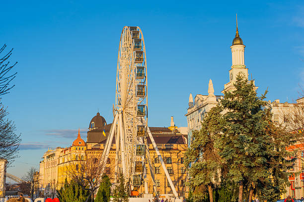 Kyiv, Ukraine - March 28, 2025: Well-lit Ferris wheel stands prominently at Kontraktova Square in Kyiv, surrounded by historic buildings and greenery, captured on a clear day with vibrant colors.