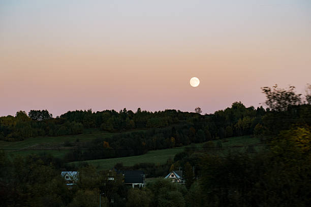 A serene countryside scene at dusk, featuring a glowing full moon rising over rolling green hills and a small village. The sky transitions from soft pink to deep blue, creating a peaceful atmosphere.