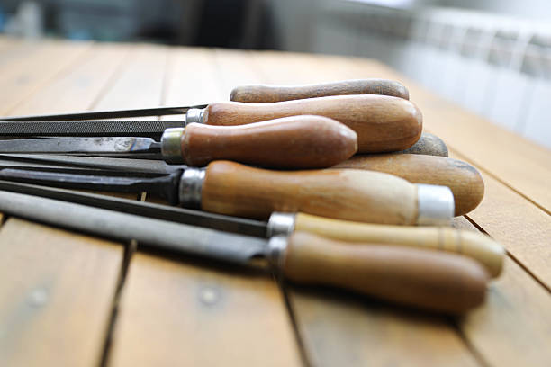 A close-up of vintage metal files with wooden handles on a rustic workbench. The focus is on the foreground file, highlighting its textured surface and worn grip, while the others fade into the background.