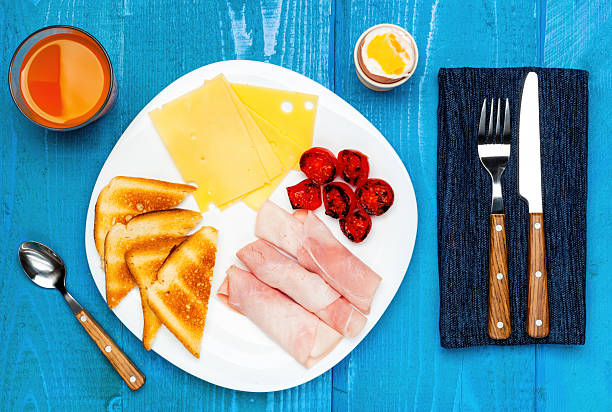 Continental breakfast is composed by ham, cheese, toast, grilled tomatoes, a soft-boiled egg and orange juice served on a white plate placed on a blue wooden table