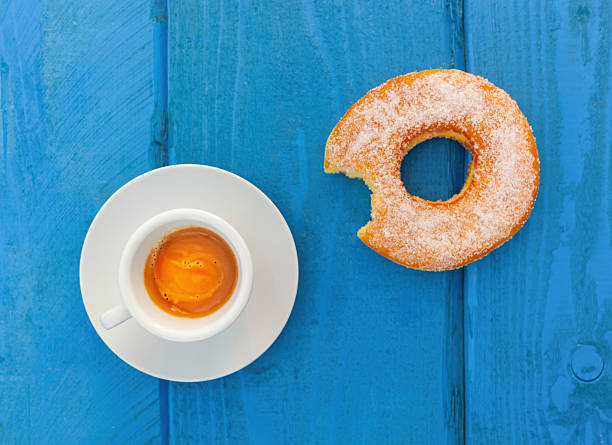 Sipping espresso coffee alongside a bitten sugared donut creates a delightful breakfast scene on a vibrant blue wooden table, inviting indulgence and warmth to the morning