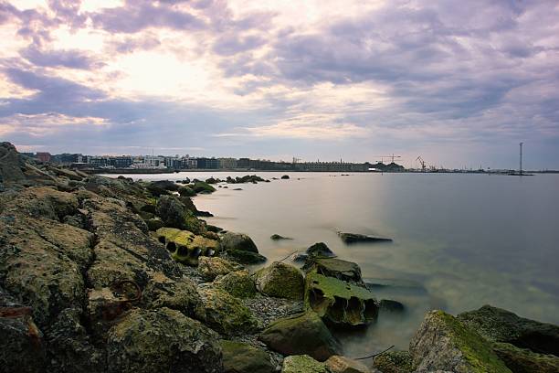 A peaceful coastal landscape that combines nature and industry. The shore is lined with large rocks covered in moss and algae, partially submerged in smooth, misty water. The dramatic sky adds depth and emotion to this atmospheric landscape.