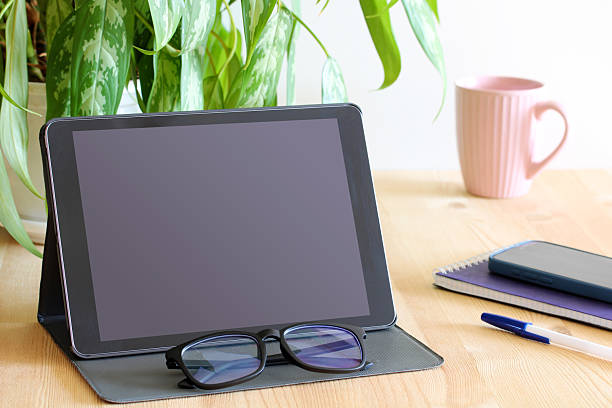 Home office desk workspace showing digital tablet with blank screen, computer eyeglasses, houseplant, coffee cup, smartphone, notebook, and pen
