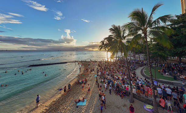 Hawaii  United States June30, 2024:This vibrant photograph captures the lively atmosphere of Waikiki Beach in Honolulu during sunset. The image showcases a large crowd gathered along the shoreline, enjoying the stunning sunset and the beach's festive ambiance. The golden light bathes the scene, highlighting the palm trees, the ocean's gentle waves, and the silhouettes of people enjoying the evening.