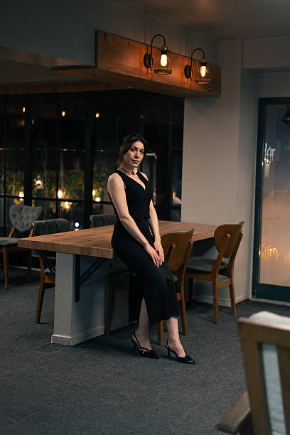 Young woman in elegant black dress sitting on a table in a modern cafe at night, looking at camera