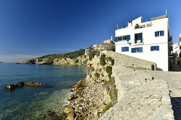 Wide-angle view of a long stretch of the southern coast of Ibiza, from the cove and beach of Platja des Cubells (at the bottom) to the verdant cliffs of Cap Llentrisca. The dazzling bright light of a Mediterranean summer noon, picturesque clouds, colourful cliffs covered with lush pine trees and a wide variety of Mediterranean shrubs and plants, scattered villas and holiday homes, a sailing boat rocking gently on crystal-clear waters. High level of detail, natural rendition, realistic feel. Developed from RAW.