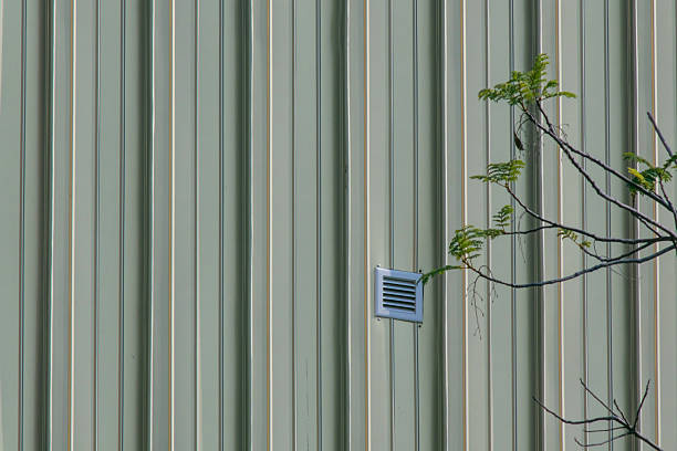 This image features a close-up of a metal wall with vertical ridges, showcasing a ventilation grille and a hint of greenery peeking from the side.