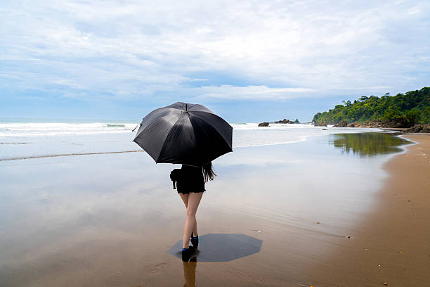 istock Slim Model Walking with a Black Umbrella on a Beach in the Pacific Ocean Surrounded by Nature in Sunlight 2208391584