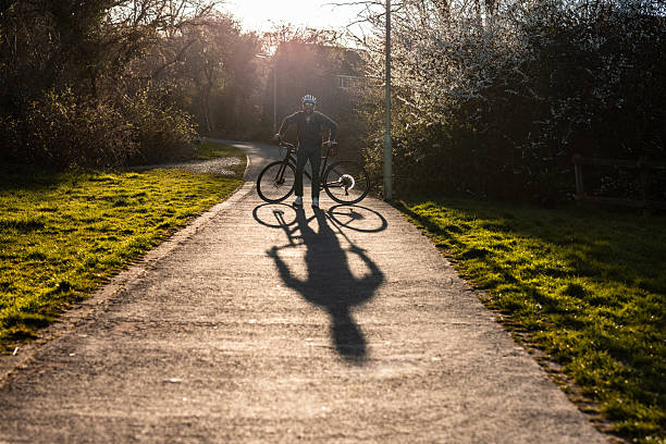 Cyclist standing with bicycle on a paved path during sunset, casting a long shadow while enjoying the warm, peaceful evening light
