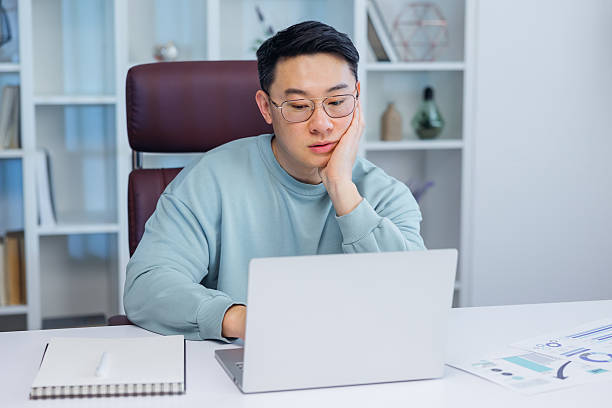 istock Bored young Asian guy sitting at desk using laptop, thinking about something during online lesson or remote work at home office. Tired man having dull distant job, feeling lazy 2208429010
