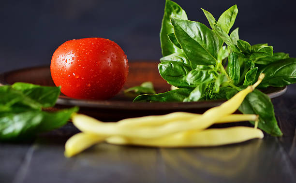 The red tomato looks appetizing among the basil leaves. Golden string beans harmoniously complement the composition. The dark wooden background emphasizes the rich colors of the vegetables.