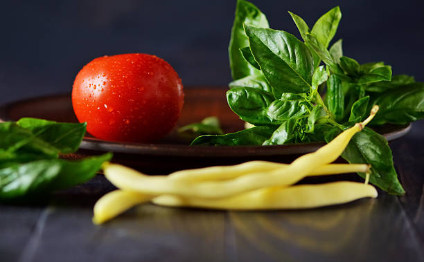 The culinary still life features a fresh tomato, aromatic basil, and bright asparagus. The texture of the vegetables stands out clearly against the black background. The contrast between the tomato and yellow beans creates visual depth.
