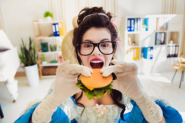 istock Young Woman in Elegant Historical Dress Playfully Eats Burger in a Modern Office Setting, Combining Past Style with Contemporary Lifestyle 2208438607