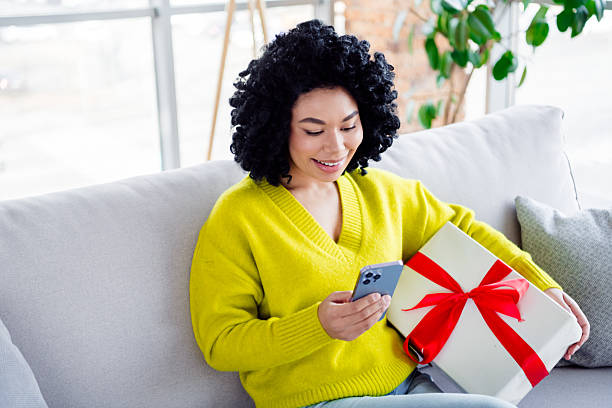 istock Young woman in bright yellow sweater relaxing on sofa while checking her smartphone and holding a gift box 2208442555