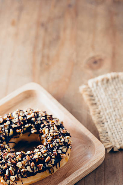 A mouthwatering chocolate donut topped with nuts sits elegantly on a wooden plate, perfect for food lovers. Ideal for cafes or dessert menus.