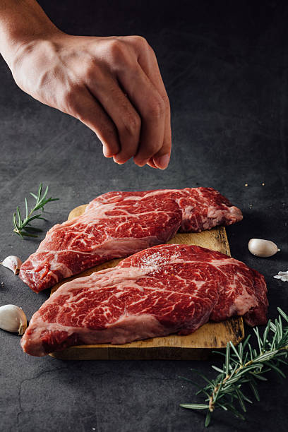 istock Hand Sprinkling Salt on Fresh Raw Meat Cuts with Herbs on Dark Background 2208443885