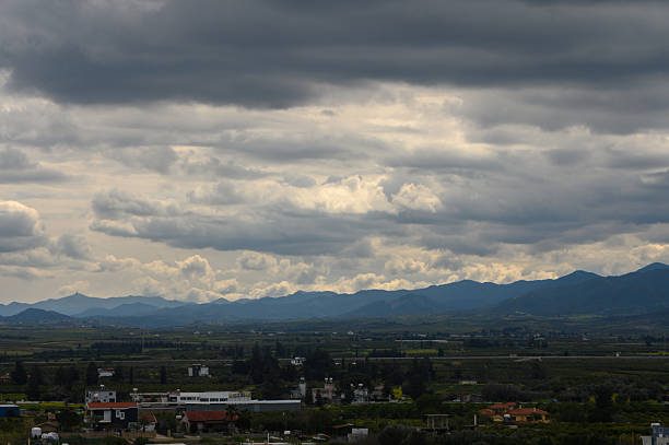 Dark clouds loom over the expansive mountain range, casting shadows across the serene fields below. The quiet landscape stretches far into the horizon, evoking a sense of calm.