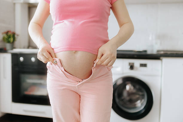istock Pregnant woman adjusting tight pants in modern kitchen setting 2208453060