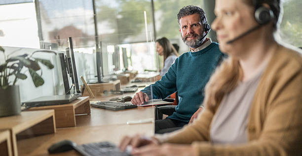 istock Mature Businessman in Open Office Wearing Headset with Colleagues in Modern Workspace 2208470330