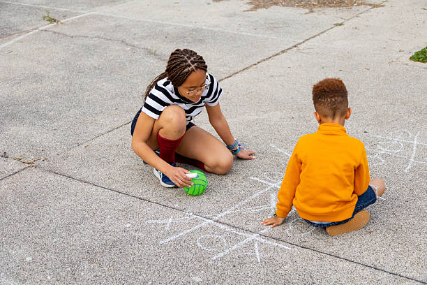 Two young children are crouching down, drawing a hopscotch grid with chalk on the pavement and preparing to play