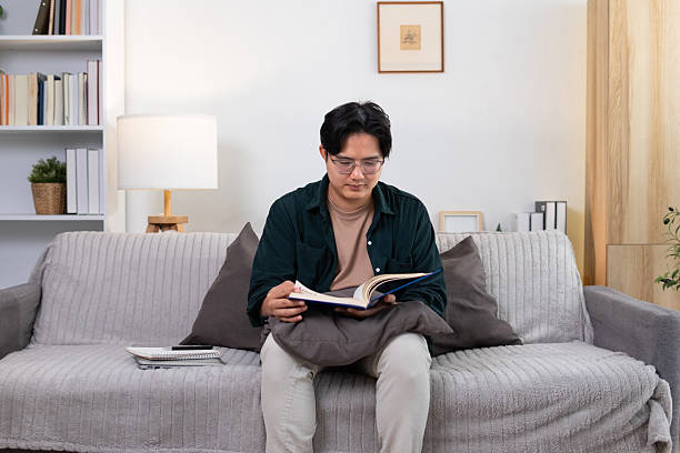 istock Inside the house, a young man sits and reads a book on a long gray sofa. 2208487226