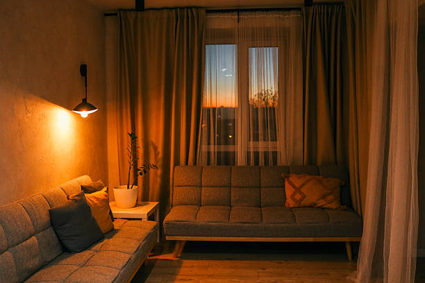 istock Cozy living room interior with warm lighting, grey sofas, decorative pillows, and a potted plant near the window during sunset 2208489873