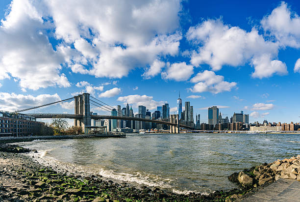 Panoramic view of the iconic Brooklyn Bridge spanning the East River with the Manhattan skyline in the background beneath a vibrant blue sky adorned with fluffy clouds.