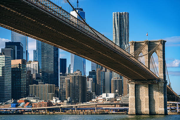 The iconic Brooklyn Bridge spans across the East River, with the impressive Manhattan skyline seen under its arch on a bright, sunny day, showcasing the architectural beauty and energy of New York City.