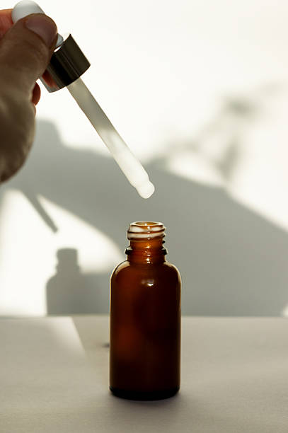 istock Brown dropper bottle without label, human hand holding pipette with white serum or cream. Cosmetic product on white background, front view, pipette in focus 2208525405