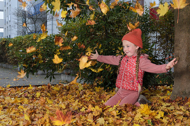 A child of six with long blonde pigtails is dressed in red and pink. She is having fun with yellow fallen maple leaves. The scene takes place in the courtyard of a city apartment building.