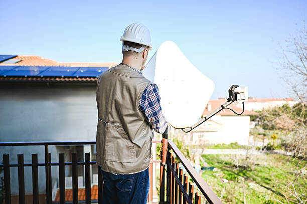 Technician installing or repairing a satellite dish on a balcony, with solar panels visible on the roof of the residential building