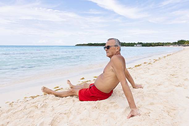 A senior man sits back on the pristine sandy shore of a tropical beach under a clear blue sky, enjoying the tranquil ocean view. The warm weather evokes relaxation and promotes summer leisure.