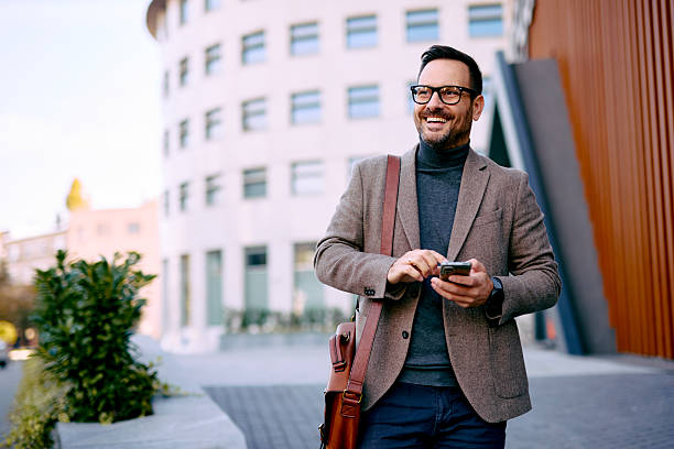istock Smiling businessman walking and using smartphone in front of office building 2208538754