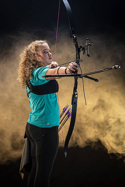 Teenage Caucasian female in a focused stance with a bow, wearing casual attire in a studio setting. Smoke and dramatic lighting highlight her concentration and skill in archery.