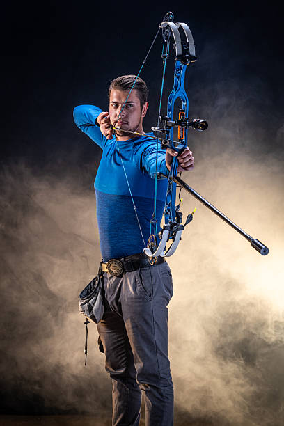 Young Caucasian male archer in a studio setting aiming an arrow with precision. Dressed in a blue shirt, he demonstrates skill and concentration. The backdrop is a dramatic blend of dark and light, enhancing the intensity.