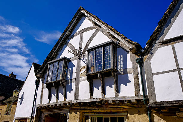 Historic street in Aylesford, Kent, featuring Tudor-style architecture. Buildings display characteristic timber-framed facades with white and dark wood detailing. Stone bases and gabled roofs add rustic charm. Large windows are symmetrically arranged, enhancing the traditional aesthetic. Street lamps line the road, which curves gently, creating a picturesque old-world charm. The early evening sky presents a soft gradient of blues and pinks, complementing the overall quaint ambiance of the scene.