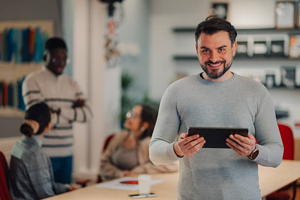 istock Smiling businessman holding tablet in modern office with colleagues working in background 2208554318
