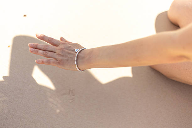 A hand rests gently on soft beach sand, adorned with a sparkling bracelet. Sunlight glints off the jewelry, highlighting the tranquil atmosphere of the seaside environment.