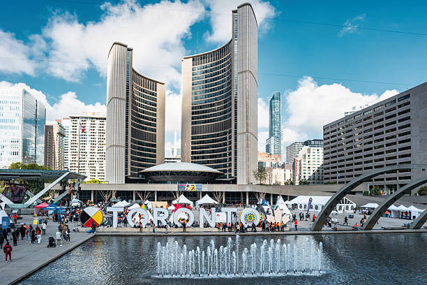 People enjoy Nathan Phillips Square in downtown Toronto, Ontario, Canada on a sunny day, with Toronto City Hall in the background.